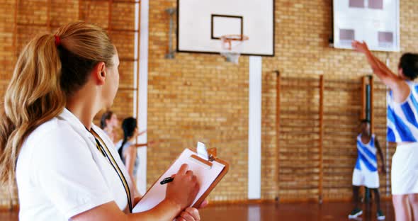 Female coach writing on clipboard while students playing in basketball court alt