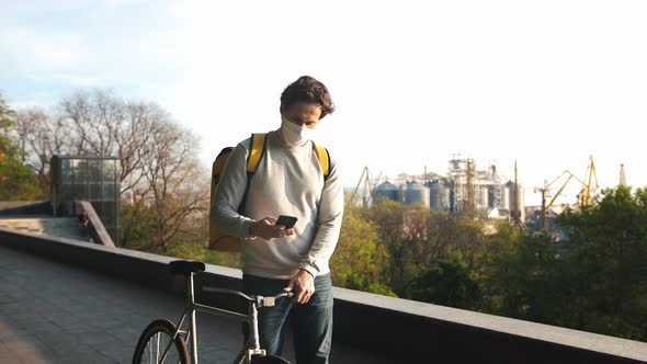 Courier with Yellow Backpack and Protective Mask Walking with Bicycle Through the City and Using alt
