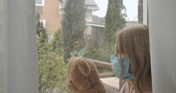 Side View of Adult Blond Caucasian Woman in Face Mask Holding Teddy Bear As Sitting on Windowsill alt