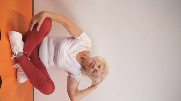 an Elderly Woman Does Morning Exercises at Home Sitting on a Fitness Mat