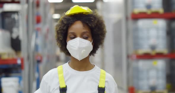Female African Worker in Safety Helmet Wearing Face Protective Mask Walking in Warehouse alt