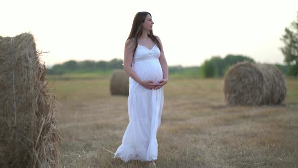 Beautiful Pregnant Woman in Wheat Field with Haystacks at Summer Day