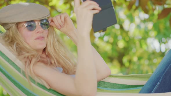 Smiling blonde woman with sunglasses using smartphone, lying relaxing on the hammock in the garden