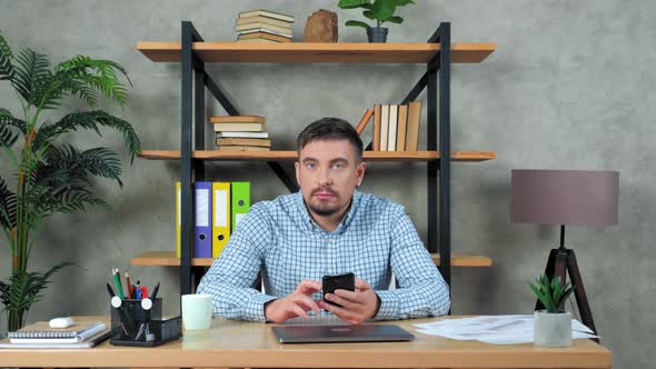 Funny businessman sitting on chair at desk in office shakes head uses smartphone alt