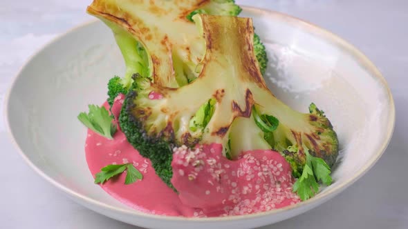 Grilled Broccoli Steak with Pink Beetroot Sauce and Hemp Seeds in White Bowl Plate is Rotating alt