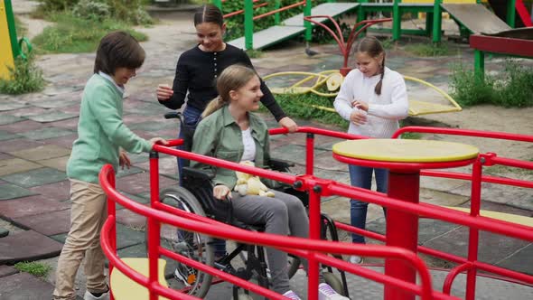 Cheerful Children Spinning Disabled Girl in Wheelchair on Merry Go Round Outdoors alt