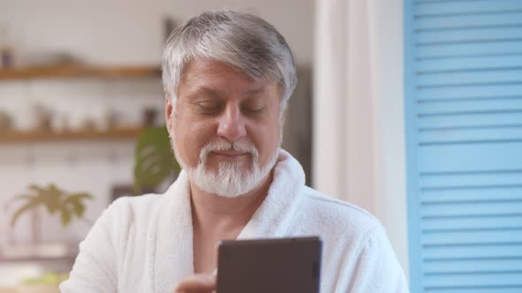Senior Man with a Gray Beard Using Digital Tablet Standing Near Window at Home alt