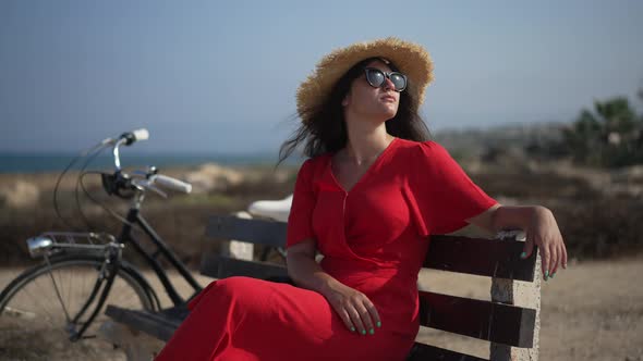 Relaxed Smiling Woman in Sunglasses Looking Away Sitting on Bench with Bike Outdoors alt