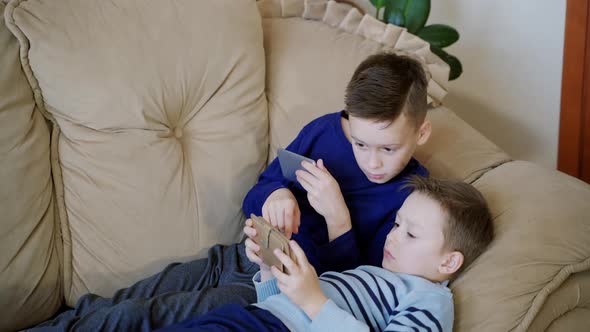 Two cute boys relaxing on the couch and playing on his smaptphones at home. alt