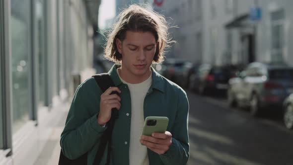 Young Man Walking on City Street and Using Cell Phone alt