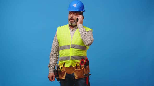 Senior Man Builder in Vest and Hardhat Using His Smartphone Against Blue Background