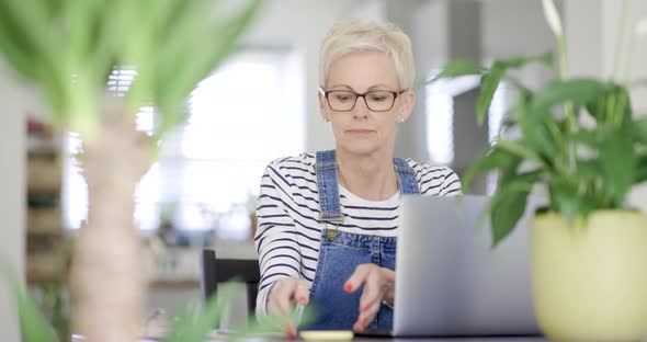 Mature woman with eyeglasses working at home on laptop alt