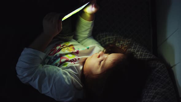 Baby Girl is Watching Video on Smartphone in Bed Before Sleeping at Evening alt
