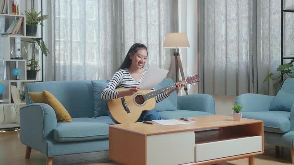 Smiling Asian Woman Composer Looking At Paper In Hand And Shaking Her Head Before Playing A Guitar alt
