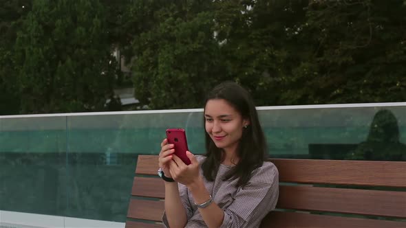 Young Beautiful Girl Sitting on a Bench in the Park, Smiling and Writing a Message on the Internet alt