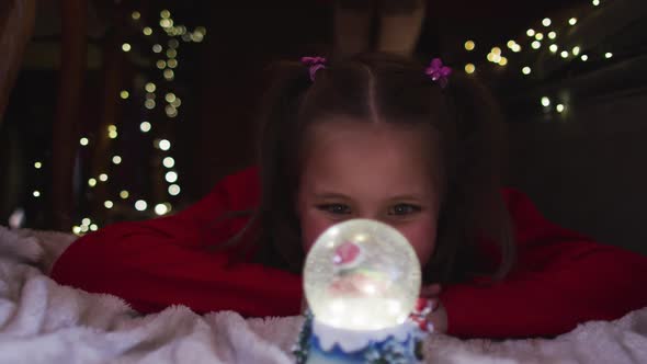 Caucasian girl smiling and looking at snow globe while lying under blanket fort during christmas alt