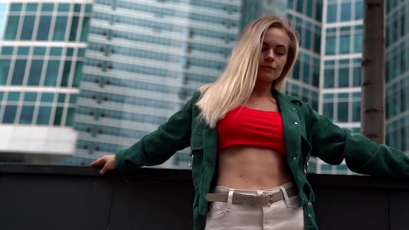 a Blonde Woman in a Red Top and Green Shirt Poses Against a Backdrop of High-rise Buildings. Bottom alt