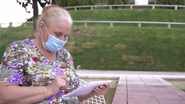 An Old Elderly Woman in a Medical Mask Uses a Tablet Computer Outdoors alt
