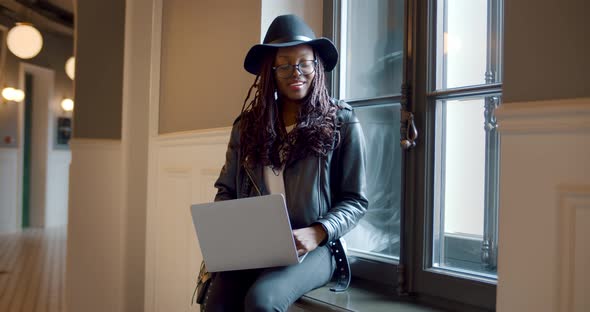 Portrait of Stylish African Woman Sitting on Windowsill Working on Laptop alt