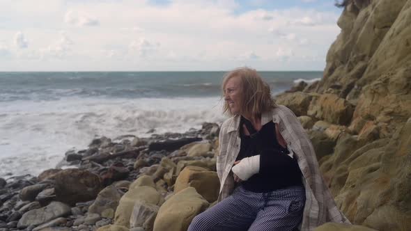 Senior Elderly Woman Relaxing Sitting on Coast of Ocean or Sea Windy Cloudy Day Suffering From Pain