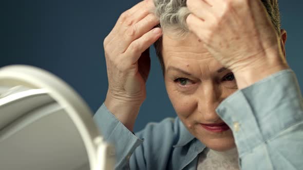 Grayhaired Elderly Woman Straightens Her Hair in Front of a Mirror Before Leaving the House alt