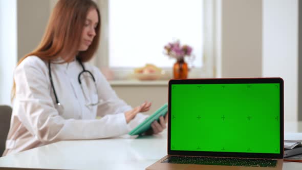 Laptop with Green Screen and Blurred Female Doctor Messaging Online on Tablet at Background alt