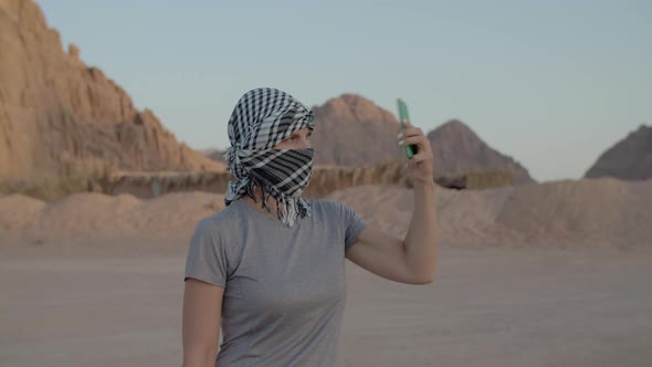 Woman in Checkered Keffiyeh Trying to Find Mobile Network Using Mobile Phone Standing in Desert alt