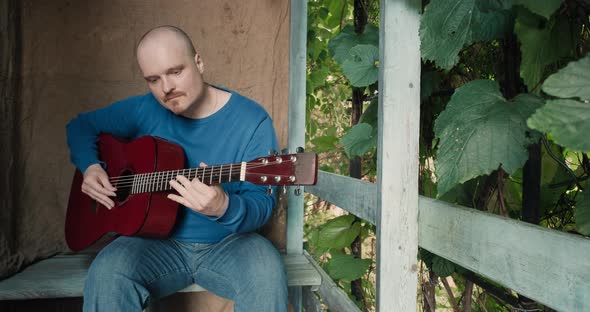 Man with an Acoustic Guitar Sits on Porch of Farmhouse He Tunes the Instrument