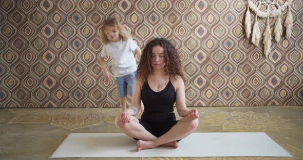 Curlyhaired Mother Meditates While Sitting on a Yoga Mat While Active Energetic Baby Daughter Jumps