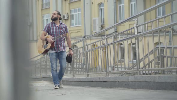 Thoughtful Caucasian Man Walking Along European City Street with Guitar and Hat. Portrait of Sad alt