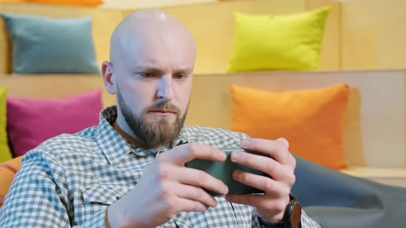 Close Up of an Young Man Sits on a Bean Bag While Playing Online Using His Phone alt
