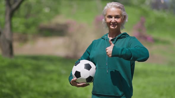 Portrait of Joyful Caucasian Senior Woman Holding Soccer Ball Showing Thumb Up Smiling alt