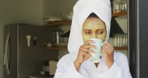 Happy african american woman with beauty mask on face, drinking coffee in kitchen alt