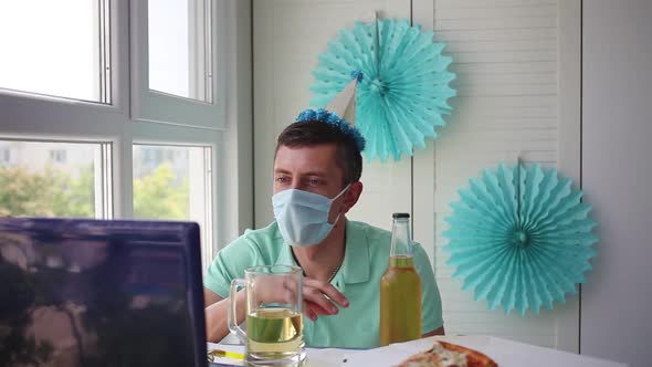 A man in a medical mask sits in front of a laptop and waves hello
