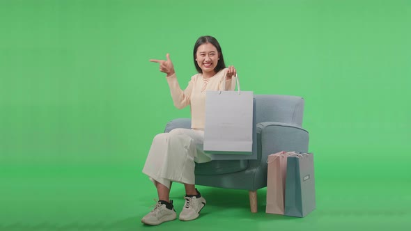 Woman Holding Shopping Bags Pointing Finger To The Side While Sitting On Sofa In Green Screen alt