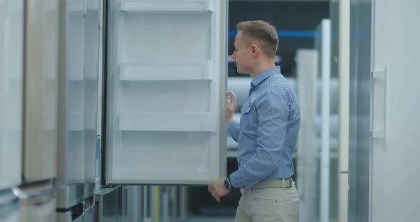The Man Open the Door of the Refrigerator in the Appliances Store and Compare with Other Models To alt