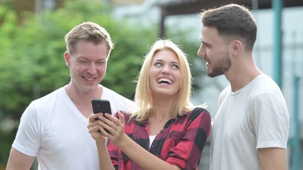 Three Happy Young Friends Smiling While Using Phone Together in the Streets Outdoors