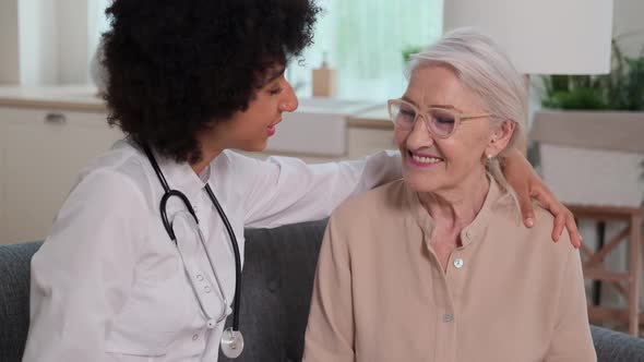 Afro American Woman Doctor Supports Senior Woman with Hand on Shoulder While Sitting on Sofa alt