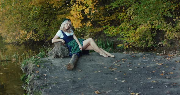 Happy Blonde in Slavic Costume Admires Autumn Nature Near River at Tree Stem alt