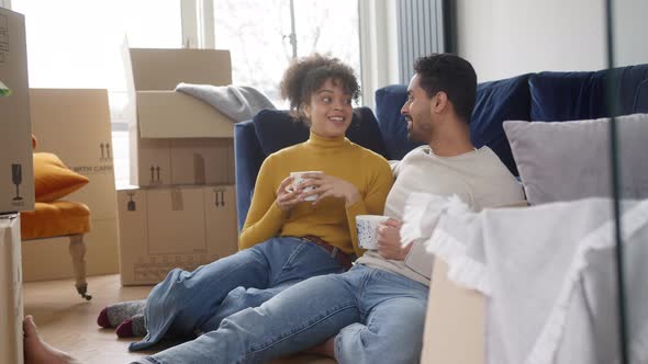 Young Couple In New Home Drink Coffee Sitting On Floor In Lounge On Moving Day With Removal Boxes