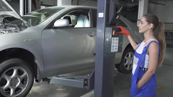 Female Young Automaster Presses a Button on a Jacking Lifting Transport Mechanism To Lower the Car alt