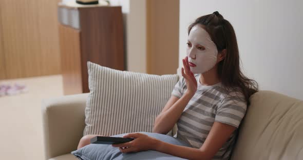 Woman apply face mask and watch TV at home alt