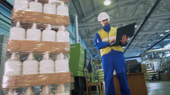 A Man Looks at a Wrapping Machine Working, Types on a Laptop alt