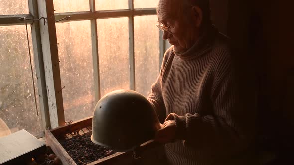 Elderly Man Glasses Picks Up Military Helmet Brushing Dust Off in House Window alt