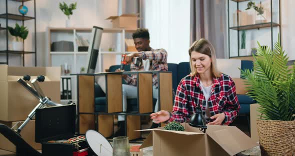 Guy Assembling Rack when His Cute Positive European Girlfriend Unpacking Boxes alt
