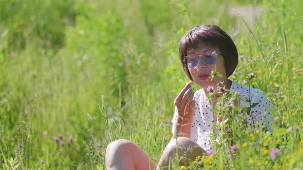 Woman in Colorful Sunglasses Enjoys Sunlight and Flower Fragrance on Grass Field alt