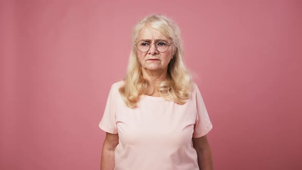 Irritated Senior Lady in Glasses Looking Around and Shushing to Camera Putting Finger on Lips Pink alt