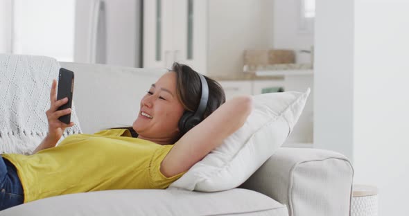 Happy asian woman wearing headphones lying on sofa, resting with smartphone at home alt