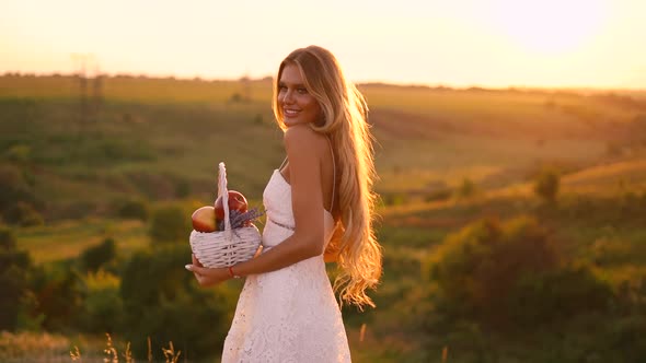Beautiful sexy blonde girl in white dress posing in a field at sunset with a basket of fruit	 alt