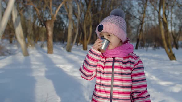 Smiling Child Kid Girl Drinking Hot Drink Tea From Cup Trying to Keep Warm in Winter Park Forest alt
